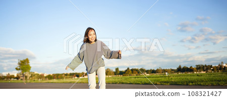 Cute asian girl riding skateboard, skating on road and smiling. Skater on cruiser longboard enjoying outdoors on sunny day Cute asian girl riding skateboard, skating on road and smiling. Skater on cruiser longboard enjoying outdoors on sunny day 108321427
