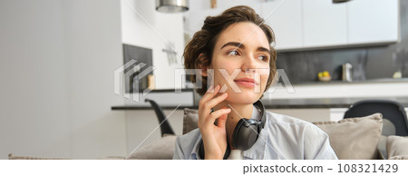 Portrait of smiling girl with headphones, looks aside, enjoys weekend at home, listens music in wireless earphones 108321429
