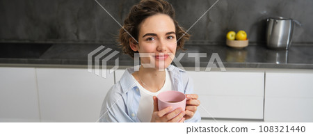 Portrait of smiling, beautiful young woman, drinking coffee in kitchen, morning magic with cuppa tea, looking tenderly Portrait of smiling, beautiful young woman, drinking coffee in kitchen, morning magic with cuppa tea, looking tenderly 108321440