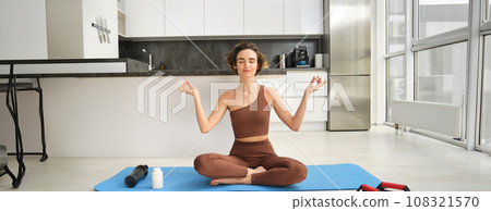 Portrait of young fitness woman, female athlete practice yoga from home, sitting in lotus pose with relaxed face, using rubber mat in kitchen for workout 108321570