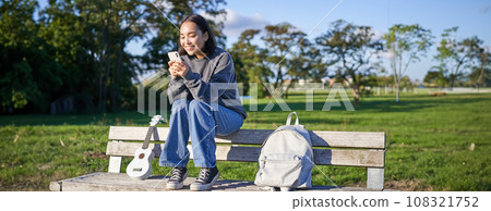 Young woman sitting in park on bench with ukulele, looking at smartphone, reading message on mobile phone and smiling 108321752