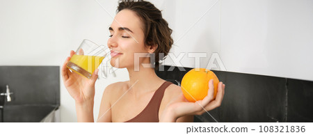 Healthy lifestyle and sport. Beautiful smiling woman, drinking fresh orange juice and holding fruit in her hand, enjoying her vitamin drink after workout at home, standing in kitchen 108321836
