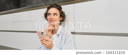 Portrait of girl with mobile phone, sits on floor at home, orders takeaway in app. Young woman with smartphone does online shopping from home, browsing websites Portrait of girl with mobile phone, sits on floor at home, orders takeaway in app. Young woman with smartphone does online shopping from home, browsing websites 108321838