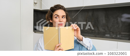 Portrait of beautiful young woman, looking intrigued, reading notes and smiling with thoughtful face, sitting at home in kitchen 108322001