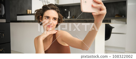 Sporty young female athlete, girl takes selfie on smartphone, workout from home and makes pictures, records video on phone during fitness exercises 108322002