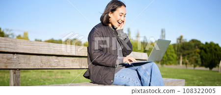 Portrait of smiling asian girl sits on bench in park, talks to friend online via laptop, video chats, using computer to study on remote Portrait of smiling asian girl sits on bench in park, talks to friend online via laptop, video chats, using computer to study on remote 108322005