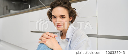 Close up portrait of young woman at home. Cute modern girl sitting on kitchen floor, smiling and looking at camera 108322043