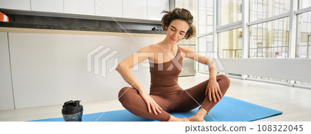 Portrait of fitness girl doing yoga on rubber mat at home, workout indoors in kitchen wearing sportswear, practice minfulness, sitting in lotus pose with water bottle next to her 108322045