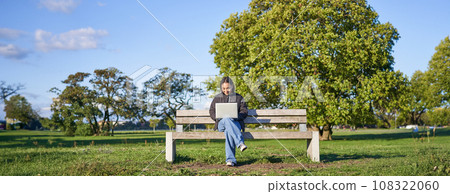 Portrait of young asian woman working on laptop, sitting on bench in park on sunny day, using her computer, studying in quite place 108322060