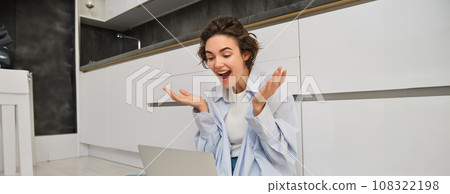 Portrait of young freelance girl connects to online meeting, video chats on laptop, sits on kitchen floor and enjoys her conversation 108322198