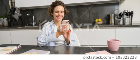 Technology and lifestyle. Young woman sits at home, uses smartphone in her kitchen and smiles Technology and lifestyle. Young woman sits at home, uses smartphone in her kitchen and smiles 108322228
