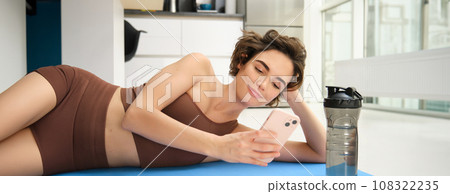 Online fitness and wellbeing. Portrait of young woman lying on yoga mat, looking at smartphone app, looking at workout tips, doing training at home in activewear Online fitness and wellbeing. Portrait of young woman lying on yoga mat, looking at smartphone app, looking at workout tips, doing training at home in activewear 108322235