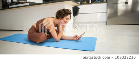 Image of young woman, fitness girl on yoga mat, workout at home, looking at her mobile phone and smiling, watches exercise videos online on smartphone app, does stretching training Image of young woman, fitness girl on yoga mat, workout at home, looking at her mobile phone and smiling, watches exercise videos online on smartphone app, does stretching training 108322236