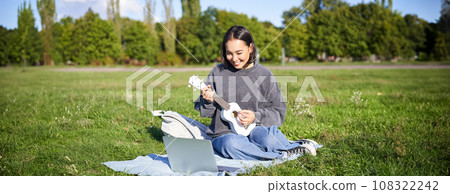 Happy asian girl plays ukulele outdoors, teaches music online with her laptop, sitting in park, playing instrument 108322242
