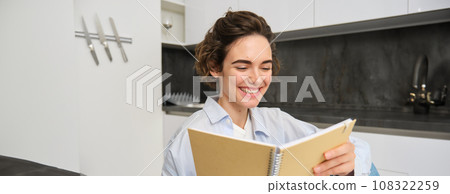 Portrait of happy brunette girl, looking at her notes, reading smth funny and laughing, sitting at home, enjoying cozy weekend Portrait of happy brunette girl, looking at her notes, reading smth funny and laughing, sitting at home, enjoying cozy weekend 108322259