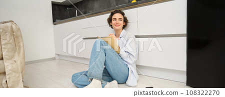 Portrait of cute brunette woman sits on floor, draws in her planner, writes diary on floor in kitchen and smiles 108322270