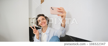 Portrait of beautiful brunette girl, takes selfie on smartphone in her kitchen, posing for photo on mobile phone Portrait of beautiful brunette girl, takes selfie on smartphone in her kitchen, posing for photo on mobile phone 108322347
