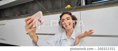 Portrait of young woman video chats, records vlog while sits on kitchen floor. Cute girl takes selfie on smartphone Portrait of young woman video chats, records vlog while sits on kitchen floor. Cute girl takes selfie on smartphone 108322352