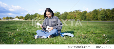 Singing asian girl playing ukulele on grass, sitting on blanket in park, relaxing outdoors on sunny day 108322429