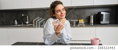 Portrait of beautiful smiling woman sitting at home with smartphone, using mobile phone app, orders groceries delivery 108322443
