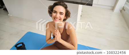 Peace, mindfulness and meditation. Young smiling sportswoman, fitness girl sits at home on floor and meditating, doing yoga, relaxing in lotus pose 108322456