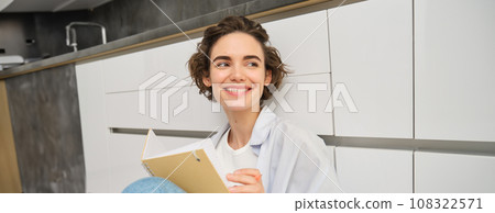 Close up portrait of young woman writing in her journal, feeling creative, sitting with planner, notebook and pen on floor, studying, doing her homework at home 108322571