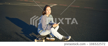 Sad asian teen girl sits on skateboard, looks upset, holds mobile phone and frowns 108322572