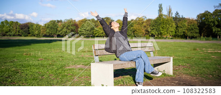 Portrait of happy asian girl feeling freedom and excitement, stretching hands while sitting on bench and smiling Portrait of happy asian girl feeling freedom and excitement, stretching hands while sitting on bench and smiling 108322583