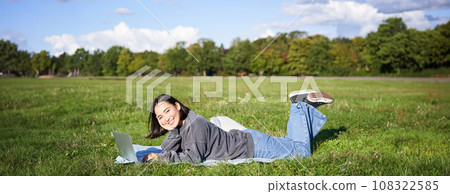 Leisure and people. Happy young woman lying in park on blanket, using laptop, relaxing outdoors and watching videos online on computer Leisure and people. Happy young woman lying in park on blanket, using laptop, relaxing outdoors and watching videos online on computer 108322585