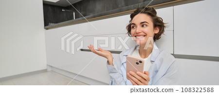 Portrait of happy young woman, shows her brand new kitchen, sits on floor, points at furniture and smiles, video chats on smartphone Portrait of happy young woman, shows her brand new kitchen, sits on floor, points at furniture and smiles, video chats on smartphone 108322588