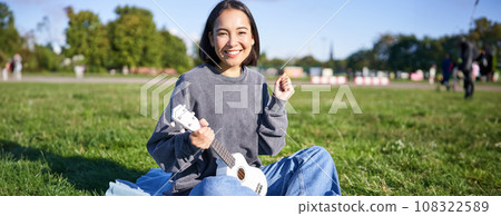 Portrait of excited asian girl, sitting with ukulele in park on green grass, looking amazed and surprised with good news Portrait of excited asian girl, sitting with ukulele in park on green grass, looking amazed and surprised with good news 108322589