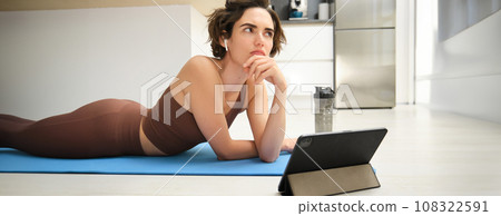Portrait of fitness girl with thoughtful face, lying on rubber yoga mat at home, listening and watching sport workout video tutorial on tablet, thinking Portrait of fitness girl with thoughtful face, lying on rubber yoga mat at home, listening and watching sport workout video tutorial on tablet, thinking 108322591