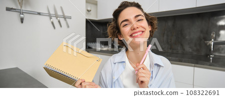 Portrait of smiling brunette girl, holds her planner, writes in notebook and looks happy, laughs, does homework, makes notes, sits at home in kitchen 108322691