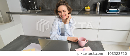 Young woman freelancer works from home, looks at laptop screen and smiles, does homework in kitchen 108322710
