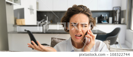 Image of woman with confused, annoyed face, talking on mobile phone while sitting at home on sofa, shrugging and looking bothered 108322744