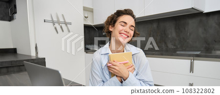 Portrait of young creative woman, holding her notebook and smiling, makes notes in her diary, writing in her planner, sitting at home in kitchen and smiling 108322802
