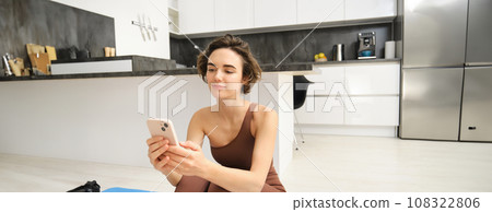 Image of beautiful woman in stylish sportswear, looking at her smartphone, sitting on yoga mat at home, workout, taking break from fitness training. Girl posting online on mobile phone Image of beautiful woman in stylish sportswear, looking at her smartphone, sitting on yoga mat at home, workout, taking break from fitness training. Girl posting online on mobile phone 108322806