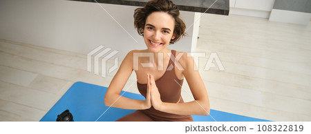 Young healthy beautiful woman in sports top and leggings practicing yoga at home, sitting in lotus position on yoga mat, meditating, smiling, relaxed, eyes closed, meditation concept Young healthy beautiful woman in sports top and leggings practicing yoga at home, sitting in lotus position on yoga mat, meditating, smiling, relaxed, eyes closed, meditation concept 108322819