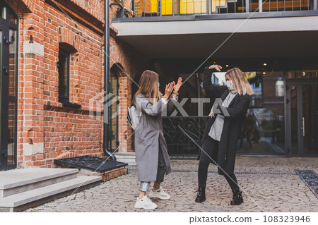 Two young stylish women in medical masks dancing and having fun while walking the street in town. Friends strolling outdoor in city Two young stylish women in medical masks dancing and having fun while walking the street in town. Friends strolling outdoor in city 108323946