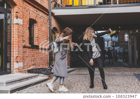 Two young stylish women in medical masks dancing and having fun while walking the street in town. Friends strolling outdoor in city Two young stylish women in medical masks dancing and having fun while walking the street in town. Friends strolling outdoor in city 108323947