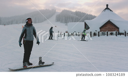 Woman enjoy winter holiday vacation on ski resort. Snowboard girl listen music, and have fun, ski slope, snow covered forest in background. Active sport. Bukovel, Carpathian mountains, Ukraine 108324189