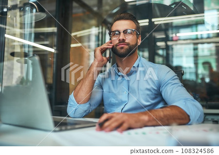 Happy to help you. Portrait of young handsome man in eyeglasses and formal wear talking on the phone with client while sitting in the office 108324586