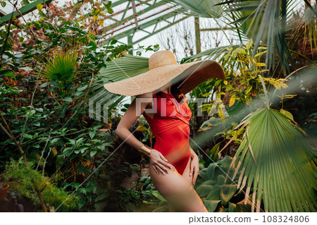 Woman in red swimsuit and with big hat standing among tropical plants in greenhouse 108324806