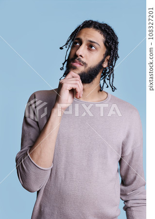 Young adult arab man with thoughtful expression looking upwards while rubbing chin. Pensive handsome arabian model with black braided hairstyle in doubt posing on blue background Young adult arab man with thoughtful expression looking upwards while rubbing chin. Pensive handsome arabian model with black braided hairstyle in doubt posing on blue background 108327201