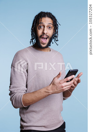 Excited arab man using smartphone and looking at camera with happy facial expression. Cheerful person in casual clothes getting online message with good news on mobile phone portrait Excited arab man using smartphone and looking at camera with happy facial expression. Cheerful person in casual clothes getting online message with good news on mobile phone portrait 108327204