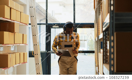 Male employee acting silly dancing in warehouse depot, feeling happy singing with music on headphones. Young cheerful man doing dance moves and being funny in storehouse, shelving. 108327239