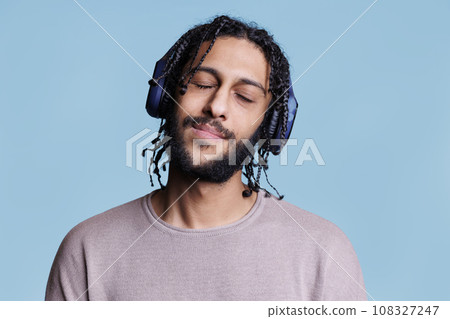 Relaxed arab man enjoying music wearing wireless headphones with closed eyes. Handsome young model in casual clothes with satisfied emotions listening to song in big earphones 108327247