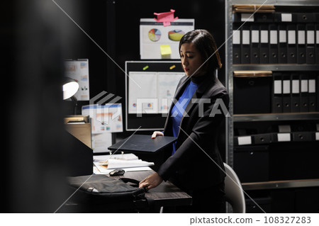 Asian woman private investigator unpacking laptop before beginning criminal investigation case. Detective in archive room office filled with criminology folders on bookshelves 108327283