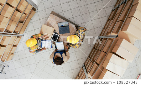 Storage room workers packing products in boxes, dping quality control and stock inventory before shipment. Young people planning merchandise distribution with packages, store delivery. Drone shot. 108327345