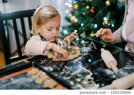 Little girl lays out cookie cutters on a piece of dough next to her mother. Cropped Little girl lays out cookie cutters on a piece of dough next to her mother. Cropped 108328906